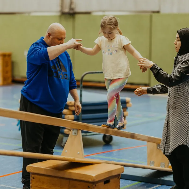 Kinder balancieren mit ihren Eltern über Bänke in der Sporthalle